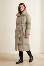 Lange Winterjacke in hellem Taupe mit Gürtel, an einem Model vor einem warmen, hellen Studiohintergrund.